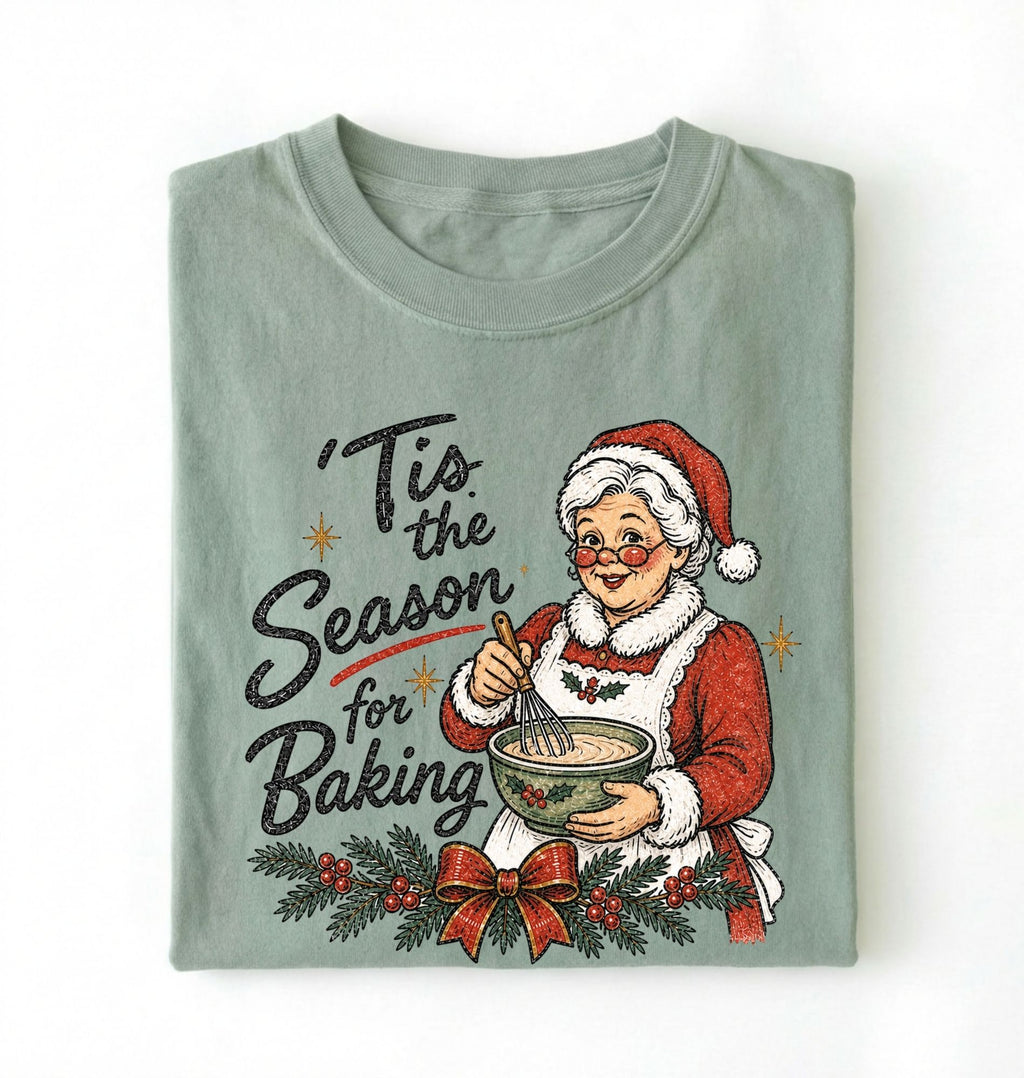A folded sage green crewneck t-shirt featuring a vintage-style Christmas graphic of an elderly woman in a Santa hat mixing batter, with the text 'Tis the Season for Baking' and a holly garland with a red bow.