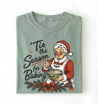 A folded sage green crewneck t-shirt featuring a vintage-style Christmas graphic of an elderly woman in a Santa hat mixing batter, with the text 'Tis the Season for Baking' and a holly garland with a red bow.