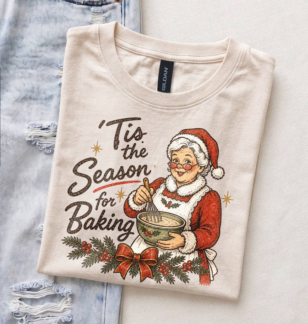 A folded cream-colored t-shirt with a vintage-style Christmas graphic of Santa Claus baking, styled flat on top of a light blue distressed denim sheet.