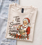 A folded cream-colored t-shirt with a vintage-style Christmas graphic of Santa Claus baking, styled flat on top of a light blue distressed denim sheet.
