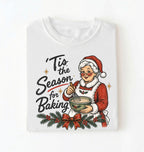 A white short-sleeve crewneck t-shirt featuring a vintage-style Christmas graphic of an elderly woman in a Santa hat and coat mixing holiday cookies, with the text 'Tis the Season for Baking'.