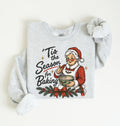 A light heather gray long-sleeve sweatshirt with a festive Christmas graphic on the front. The graphic features the text "'Tis the Season for Baking" alongside an illustration of a smiling Santa Claus in a red suit mixing ingredients in a bowl, decorated with a red bow and green pine branches.
