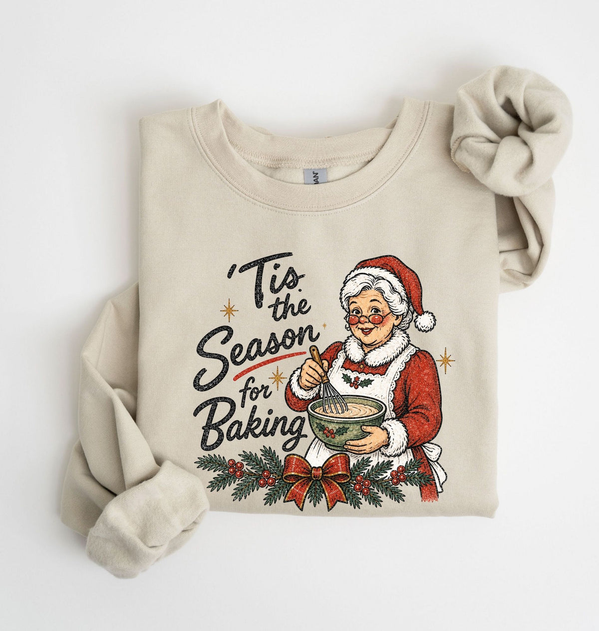 A beige crewneck sweatshirt featuring a festive Christmas graphic of an elderly woman in a Santa hat baking, with the text 'Tis the Season for Baking' and holiday greenery. The sweatshirt is styled flat on a white background with one sleeve rolled up and the other draped.