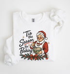 A white long-sleeve sweatshirt with a festive Christmas graphic on the front, featuring the text 'Tis the Season for Baking' and an illustration of an elderly woman in a Santa hat mixing cookies. The sweatshirt is styled flat on a clean white background with a decorative fabric loop on the right sleeve.