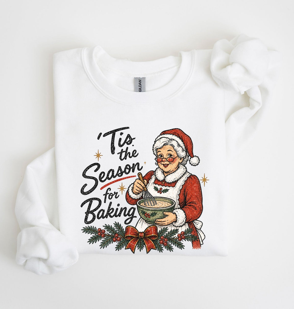 A white long-sleeve sweatshirt with a festive Christmas graphic on the front, featuring the text 'Tis the Season for Baking' and an illustration of an elderly woman in a Santa hat mixing cookies. The sweatshirt is styled flat on a clean white background with a decorative fabric loop on the right sleeve.