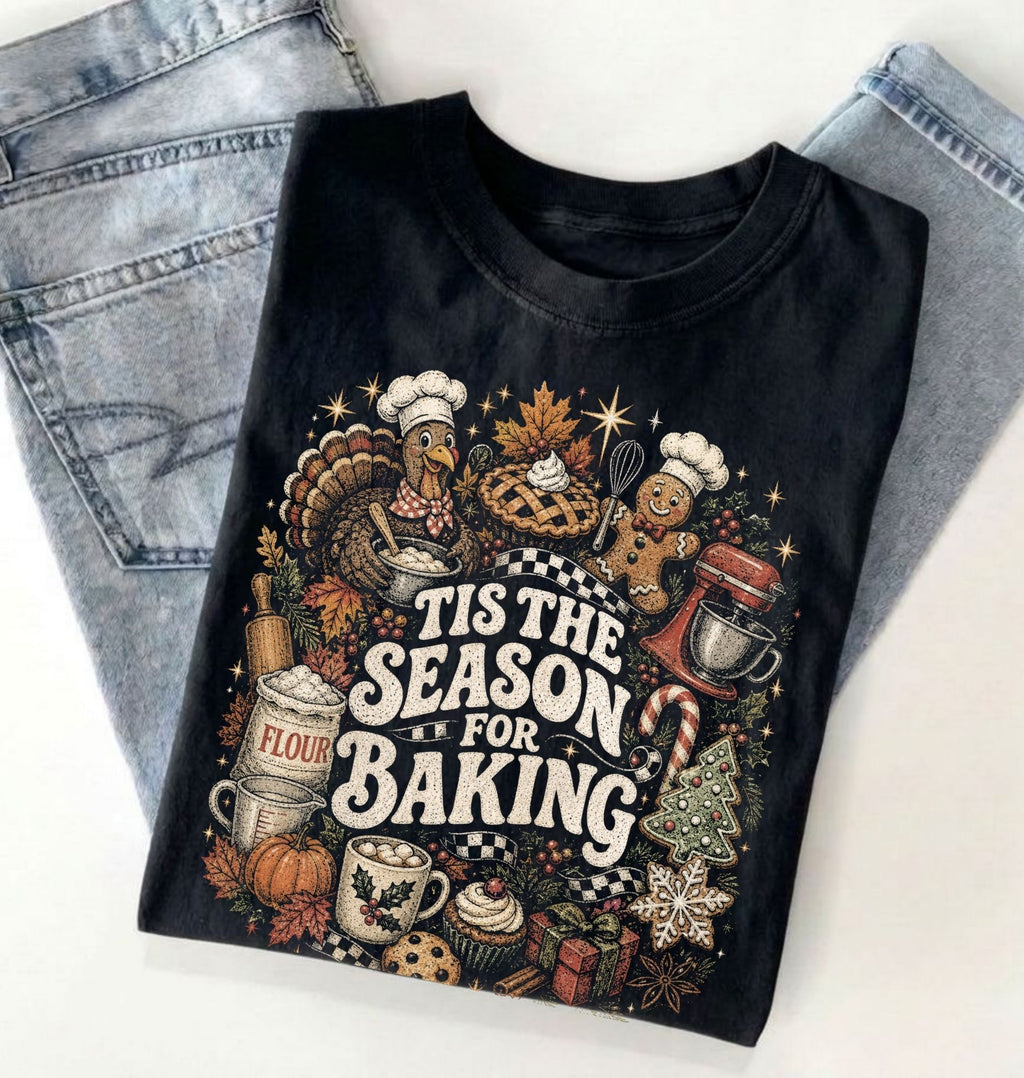 A folded black graphic T-shirt with a colorful Thanksgiving baking illustration and the text 'TIS THE SEASON FOR BAKING,' styled on top of light blue jeans against a white background.