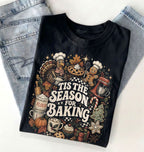 A folded black graphic T-shirt with a colorful Thanksgiving baking illustration and the text 'TIS THE SEASON FOR BAKING,' styled on top of light blue jeans against a white background.