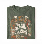 A folded dark olive green t-shirt with a colorful 'Tis the Season for Baking' graphic print featuring illustrations of a turkey, gingerbread man, stand mixer, flour sack, and cupcakes on a white background.