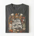 A folded dark gray t-shirt with a detailed holiday baking graphic on the front, featuring illustrations of a turkey, gingerbread man, pie, flour sack, and the text 'TIS THE SEASON FOR BAKING' on a white background.