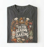 A folded dark gray t-shirt with a detailed holiday baking graphic on the front, featuring illustrations of a turkey, gingerbread man, pie, flour sack, and the text 'TIS THE SEASON FOR BAKING' on a white background.