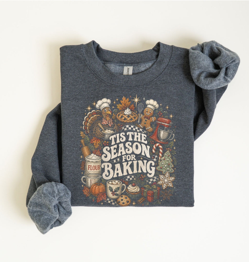 A folded dark heather gray sweatshirt with rolled sleeves, featuring a colorful graphic print that reads 'Tis the Season for Baking' surrounded by baking and autumn-themed illustrations, laid flat on a light cream background.