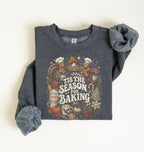 A folded dark heather gray sweatshirt with rolled sleeves, featuring a colorful graphic print that reads 'Tis the Season for Baking' surrounded by baking and autumn-themed illustrations, laid flat on a light cream background.