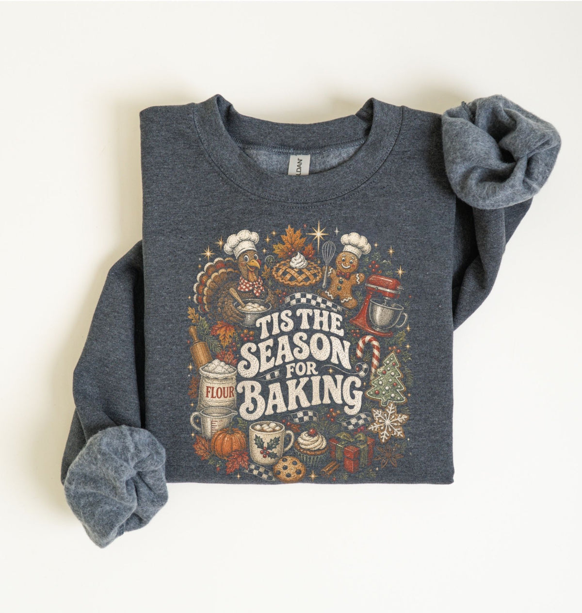 A folded dark heather gray sweatshirt with rolled sleeves, featuring a colorful graphic print that reads 'Tis the Season for Baking' surrounded by baking and autumn-themed illustrations, laid flat on a light cream background.