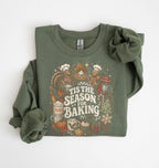 An olive green long-sleeve crewneck sweatshirt with a festive holiday baking graphic on the front, styled flat on a white background with one sleeve rolled up.