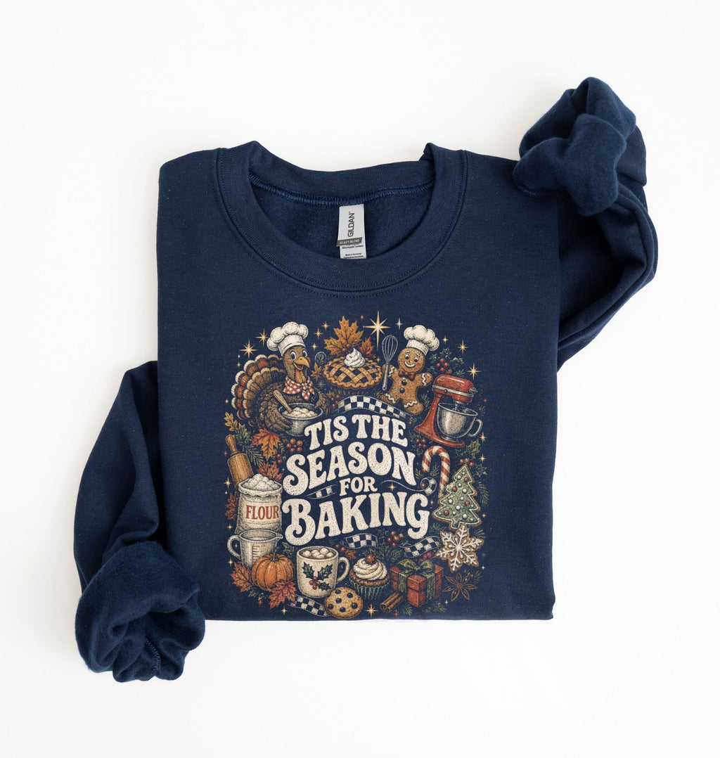 A navy blue crewneck sweatshirt with a festive 'Tis the Season for Baking' graphic print, shown folded on a white background with one sleeve rolled up.