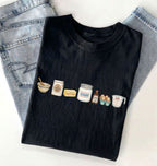 A black short-sleeve T-shirt laid flat on a white surface, styled with folded light-wash jeans behind it. The shirt has a small illustrated row of kitchen ingredients and utensils printed across the chest, including a bowl, spoon, flour sack, butter, sugar jar, vanilla bottle, eggs, and measuring cup.