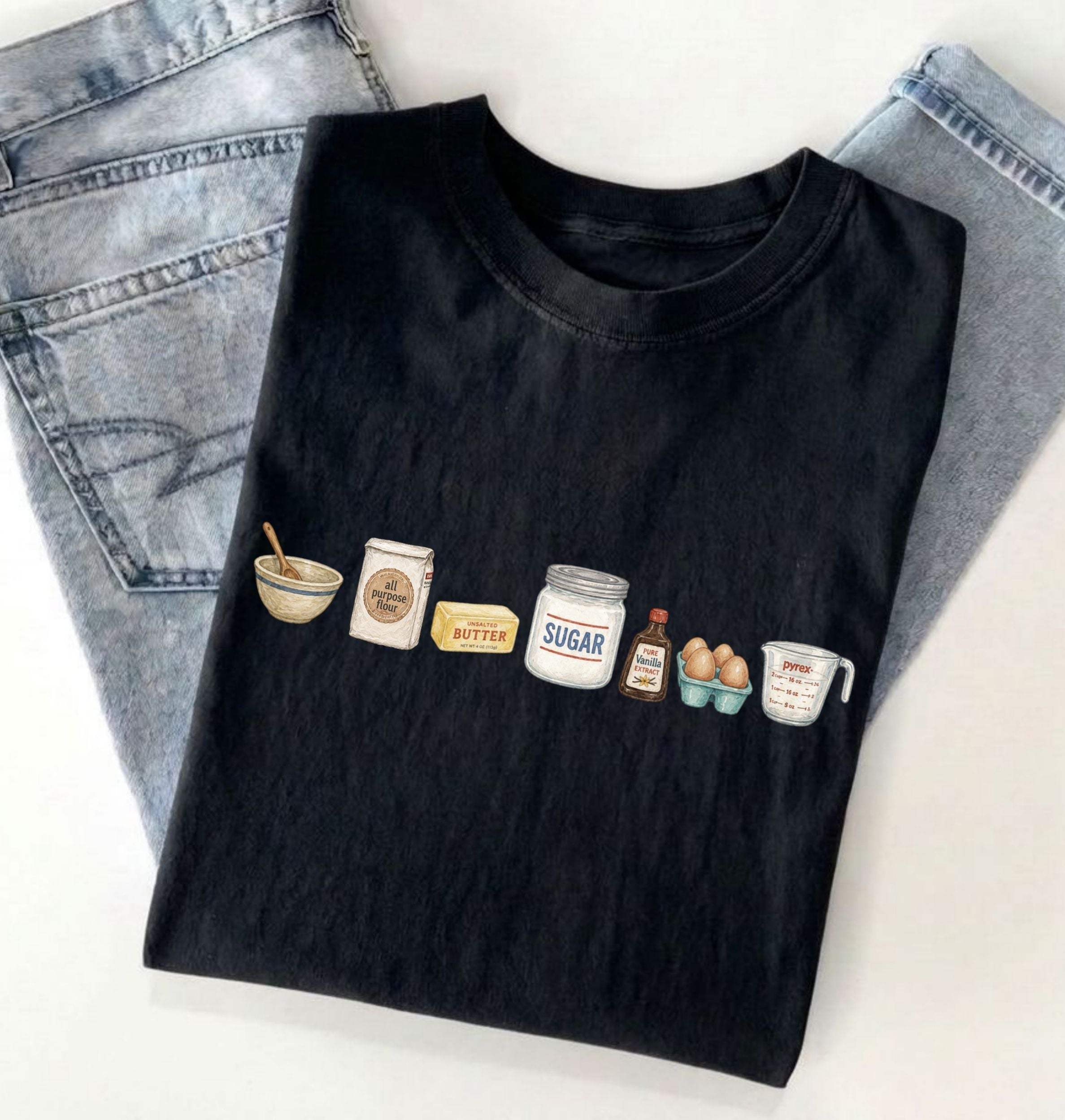 A black short-sleeve T-shirt laid flat on a white surface, styled with folded light-wash jeans behind it. The shirt has a small illustrated row of kitchen ingredients and utensils printed across the chest, including a bowl, spoon, flour sack, butter, sugar jar, vanilla bottle, eggs, and measuring cup.