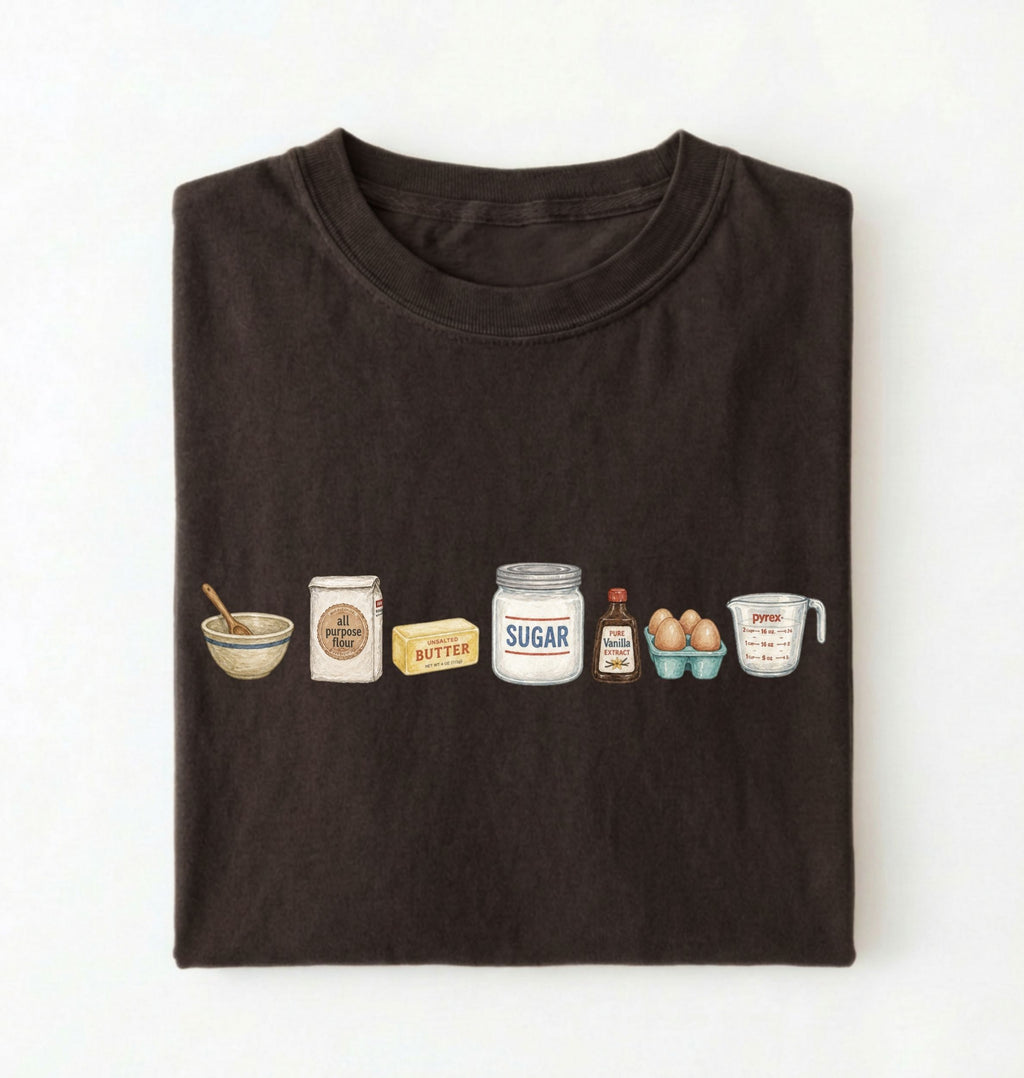 A dark brown folded t-shirt on a white background, featuring a graphic print of baking ingredients including a bowl, flour, butter, sugar, vanilla, and eggs.