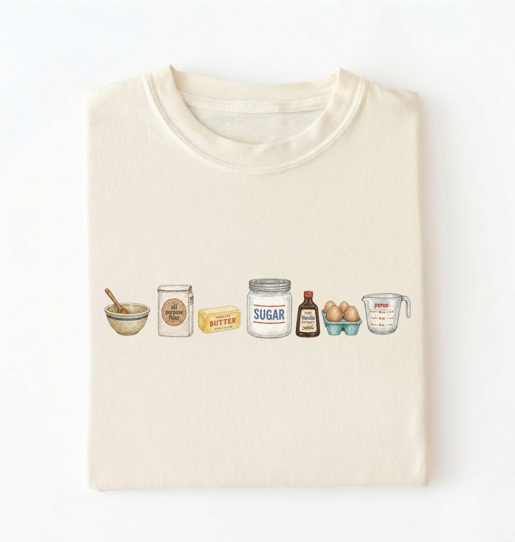 A cream-colored t-shirt folded neatly on a white background, featuring a watercolor-style graphic of baking ingredients including a bowl, flour jar, butter, sugar jar, vanilla bottle, eggs, and a measuring cup.