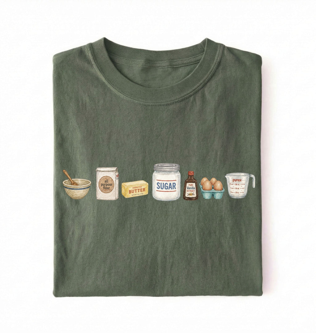 A folded dark olive green t-shirt on a white background, featuring a graphic print of baking ingredients including a bowl, flour, butter, sugar, vanilla, and eggs.