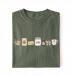 A folded dark olive green t-shirt on a white background, featuring a graphic print of baking ingredients including a bowl, flour, butter, sugar, vanilla, and eggs.
