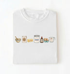 A white crewneck t-shirt with a watercolor-style graphic of baking ingredients (bowl, flour jar, butter, sugar jar, vanilla bottle, eggs, and measuring cup) across the chest, displayed flat against a white background.