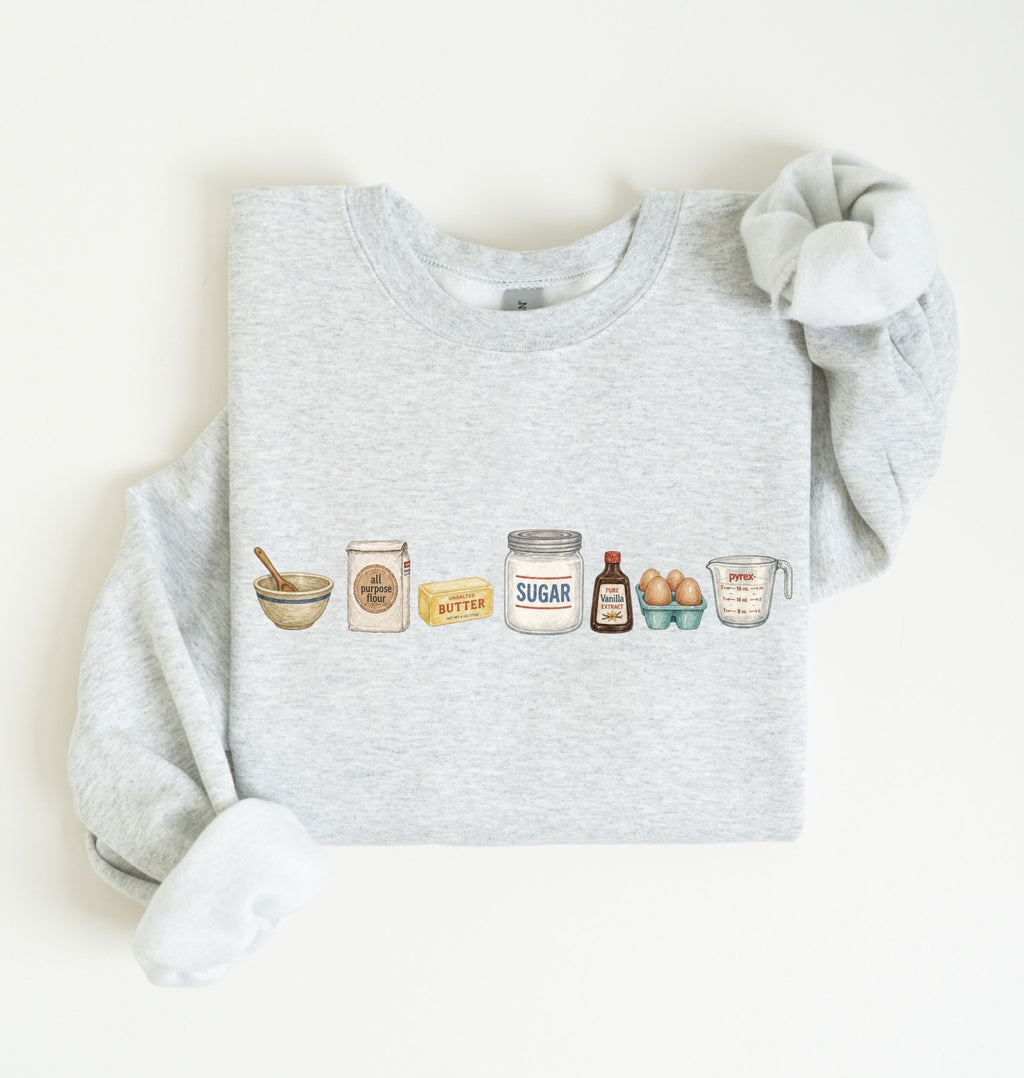 A light heather gray sweatshirt with long sleeves, laid flat on a white background. The front of the sweatshirt features a horizontal graphic illustration of baking ingredients, including a bowl, flour sack, butter, sugar jar, vanilla bottle, eggs, and a measuring cup.