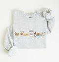 A light heather gray sweatshirt with long sleeves, laid flat on a white background. The front of the sweatshirt features a horizontal graphic illustration of baking ingredients, including a bowl, flour sack, butter, sugar jar, vanilla bottle, eggs, and a measuring cup.