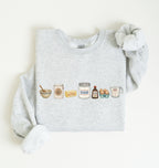 A light heather gray sweatshirt with long sleeves, laid flat on a white background. The front of the sweatshirt features a horizontal graphic illustration of baking ingredients, including a bowl, flour sack, butter, sugar jar, vanilla bottle, eggs, and a measuring cup.