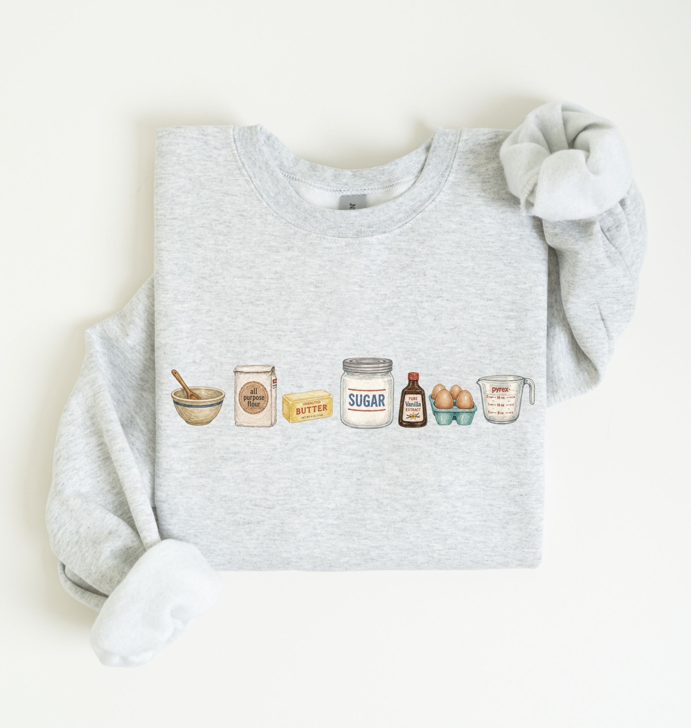 A light heather gray sweatshirt with long sleeves, laid flat on a white background. The front of the sweatshirt features a horizontal graphic illustration of baking ingredients, including a bowl, flour sack, butter, sugar jar, vanilla bottle, eggs, and a measuring cup.