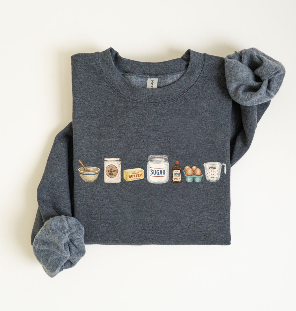 A dark heather gray crewneck sweatshirt with rolled-up sleeves, featuring a graphic print of baking ingredients (bowl, flour sack, butter, sugar jar, eggs, and measuring cup) across the chest, laid flat on a white background.