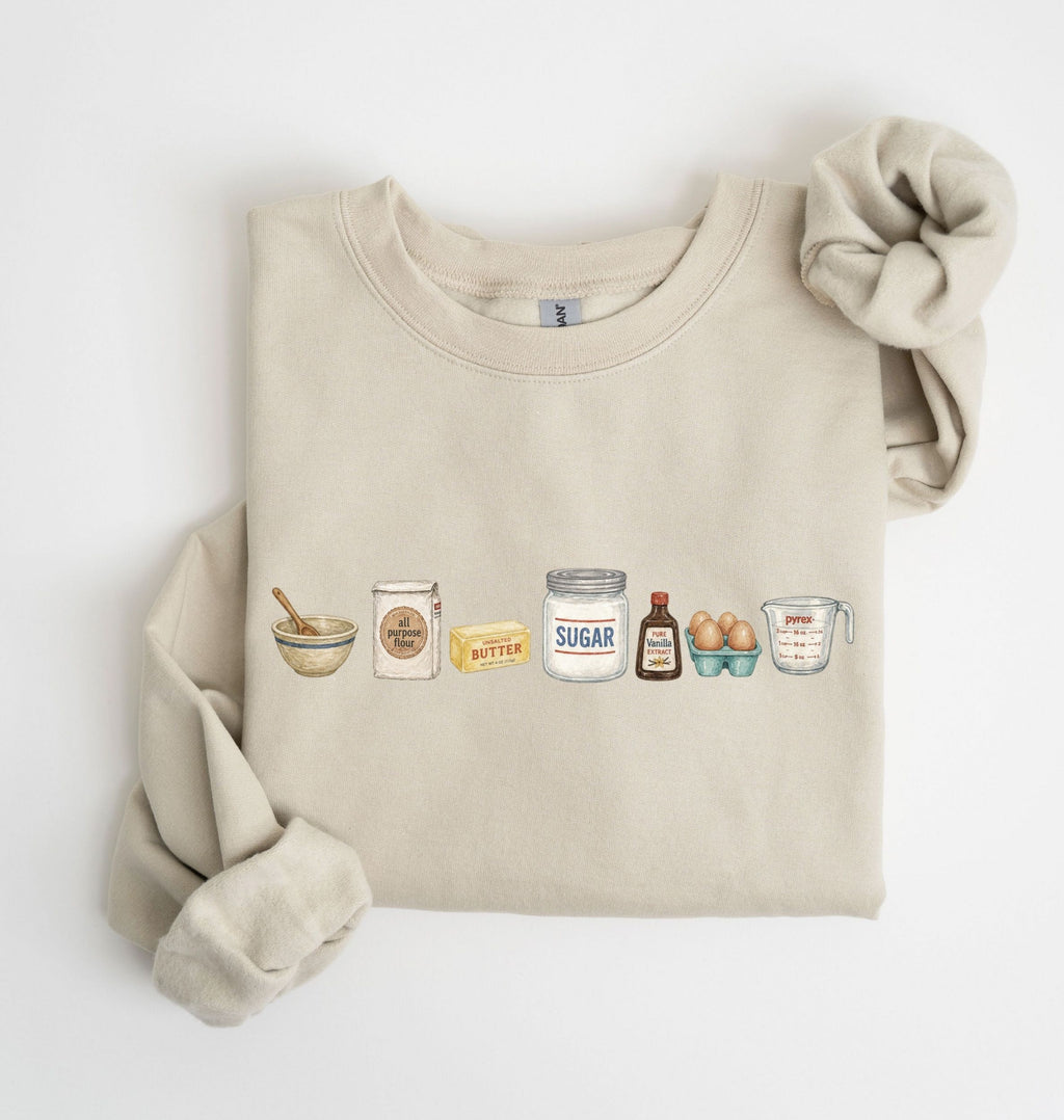 A beige crewneck sweatshirt with a graphic of baking ingredients (bowl, flour, butter, sugar, vanilla, eggs, measuring cup) printed on the chest, styled flat on a white background with a matching fabric scrunchie.