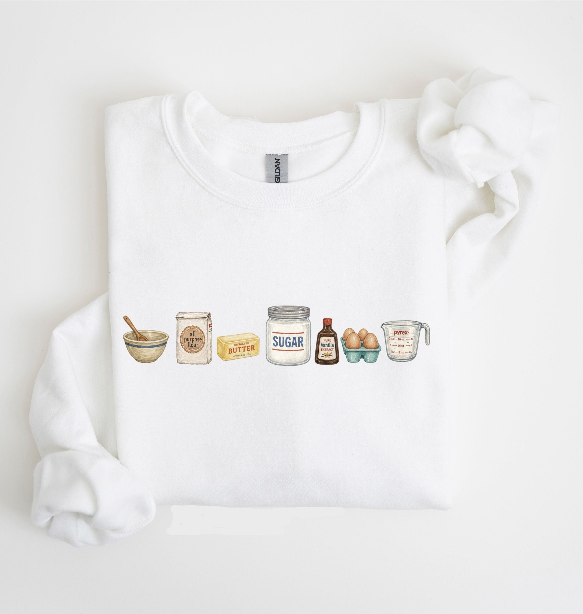 A white crewneck sweatshirt featuring a graphic print of various baking ingredients (bowl, flour, butter, sugar, vanilla, eggs, measuring cup) arranged in a row, displayed on a clean white background.