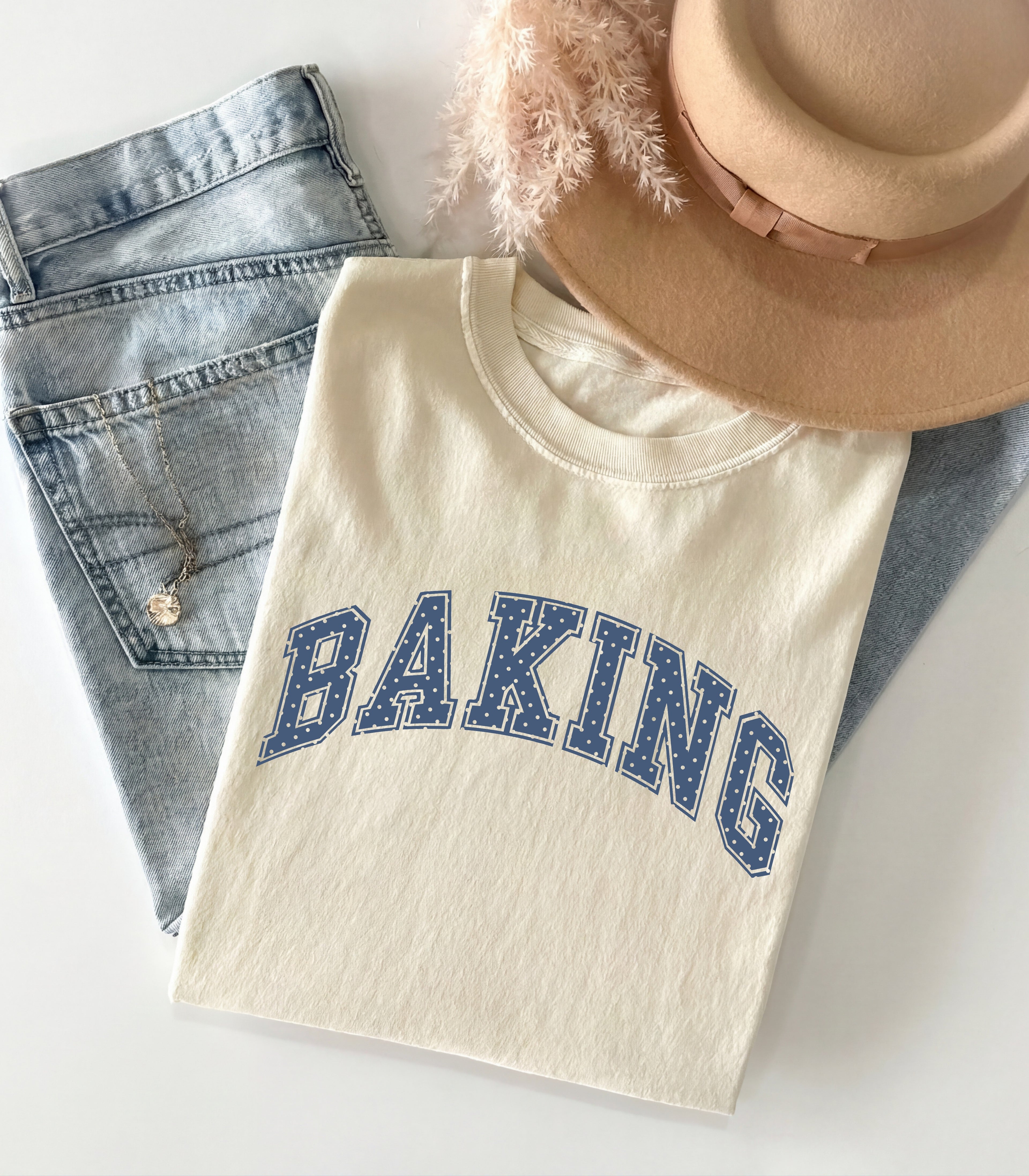 An off-white crewneck t-shirt with 'BAKING' printed in blue varsity letters, styled in a flat lay with light-wash denim jeans, a beige wide-brim hat, and a fluffy beige feather on a white background.