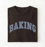 A folded dark brown t-shirt with the word 'BAKING' printed across the chest in arched varsity-style letters filled with a light blue dotted pattern, set against a plain white background.
