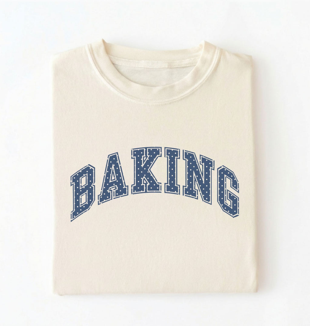 An cream folded t-shirt with 'BAKING' printed in navy blue varsity-style block letters with white dotted outlines across the chest, set against a plain white background.