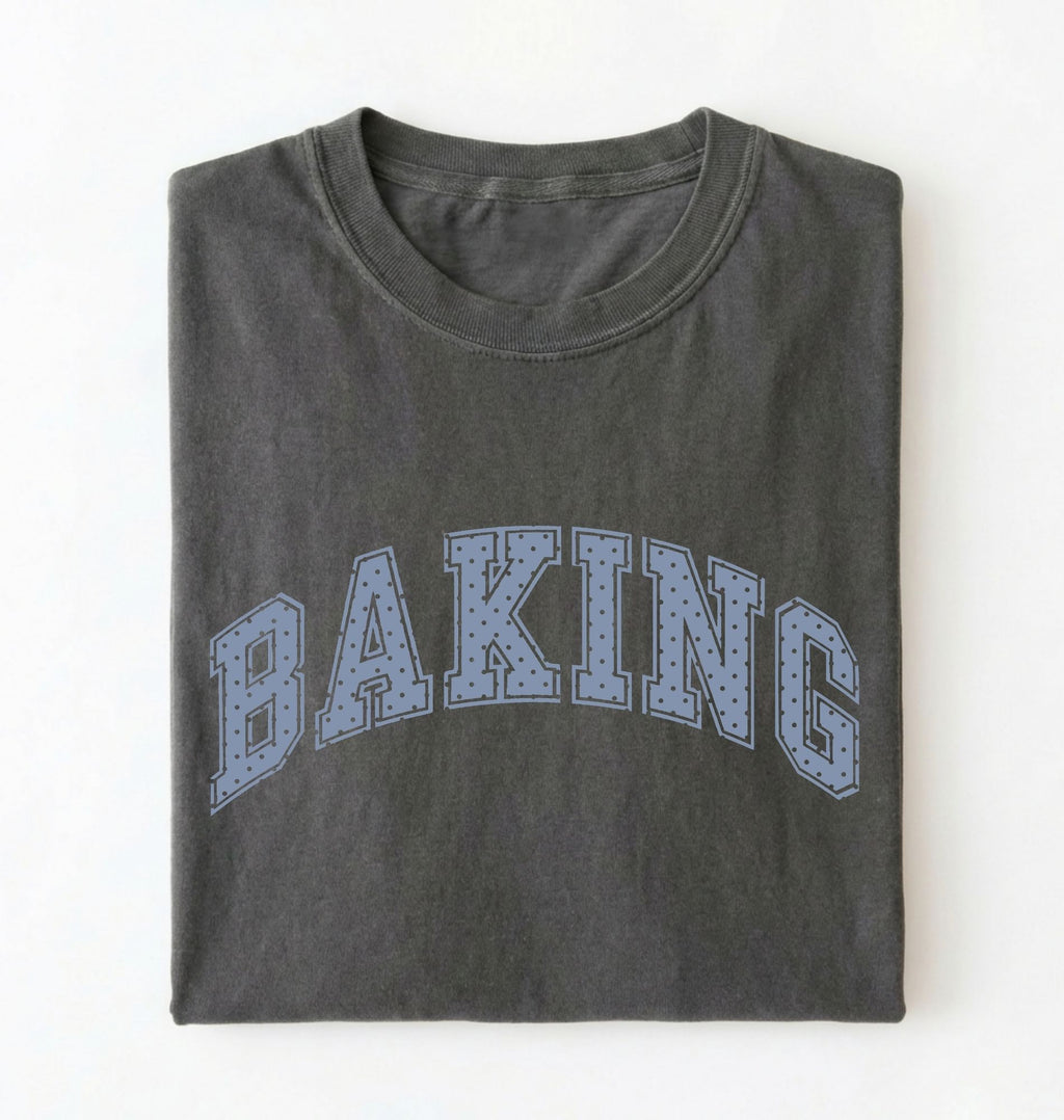 A dark gray folded t-shirt with the word 'BAKING' printed across the front in a light blue dotted collegiate-style font, shown against a plain white background.