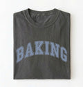 A dark gray folded t-shirt with the word 'BAKING' printed across the front in a light blue dotted collegiate-style font, shown against a plain white background.