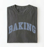 A dark gray folded t-shirt with the word 'BAKING' printed across the front in a light blue dotted collegiate-style font, shown against a plain white background.