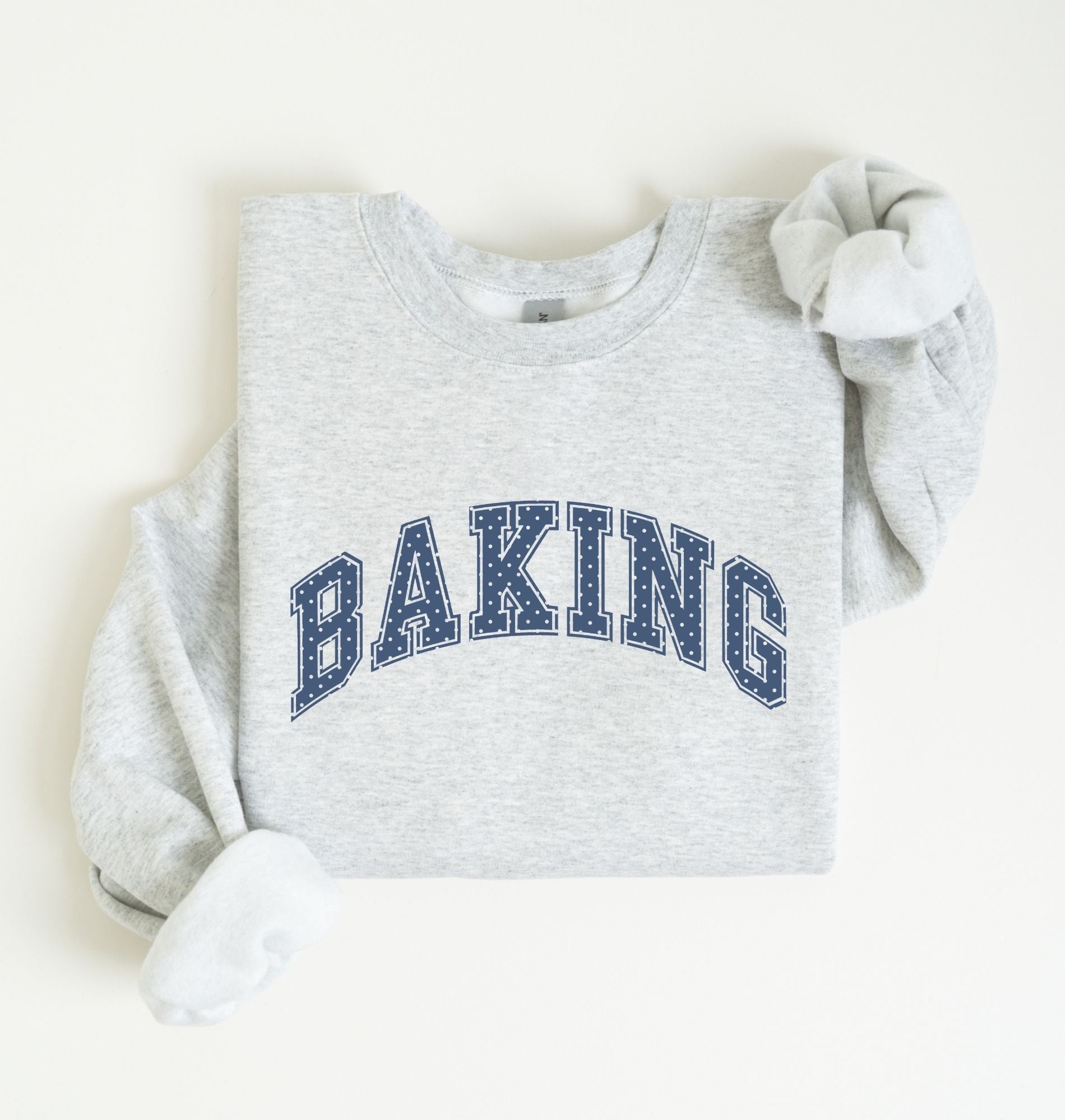 A light heather gray crewneck sweatshirt with 'BAKING' printed in navy blue varsity-style block letters across the chest, styled flat on a light cream background.