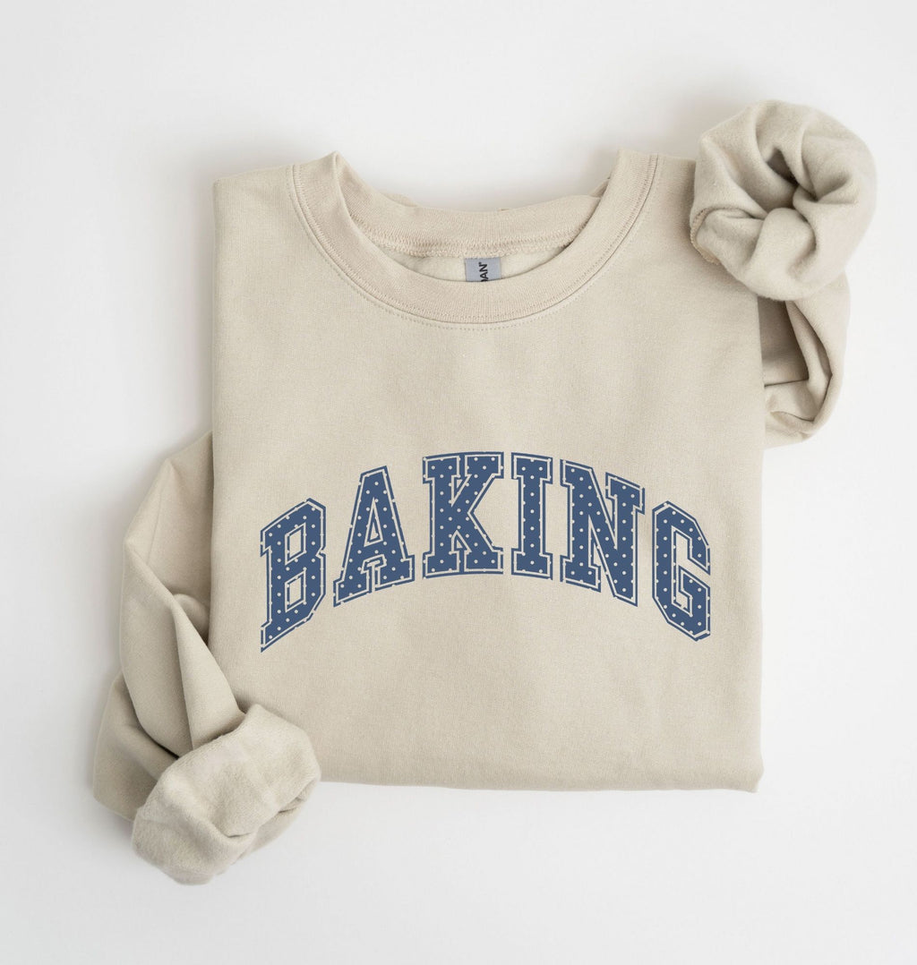 A folded cream-colored sweatshirt with 'BAKING' printed in blue varsity-style letters across the chest, styled with rolled sleeves on a white background.