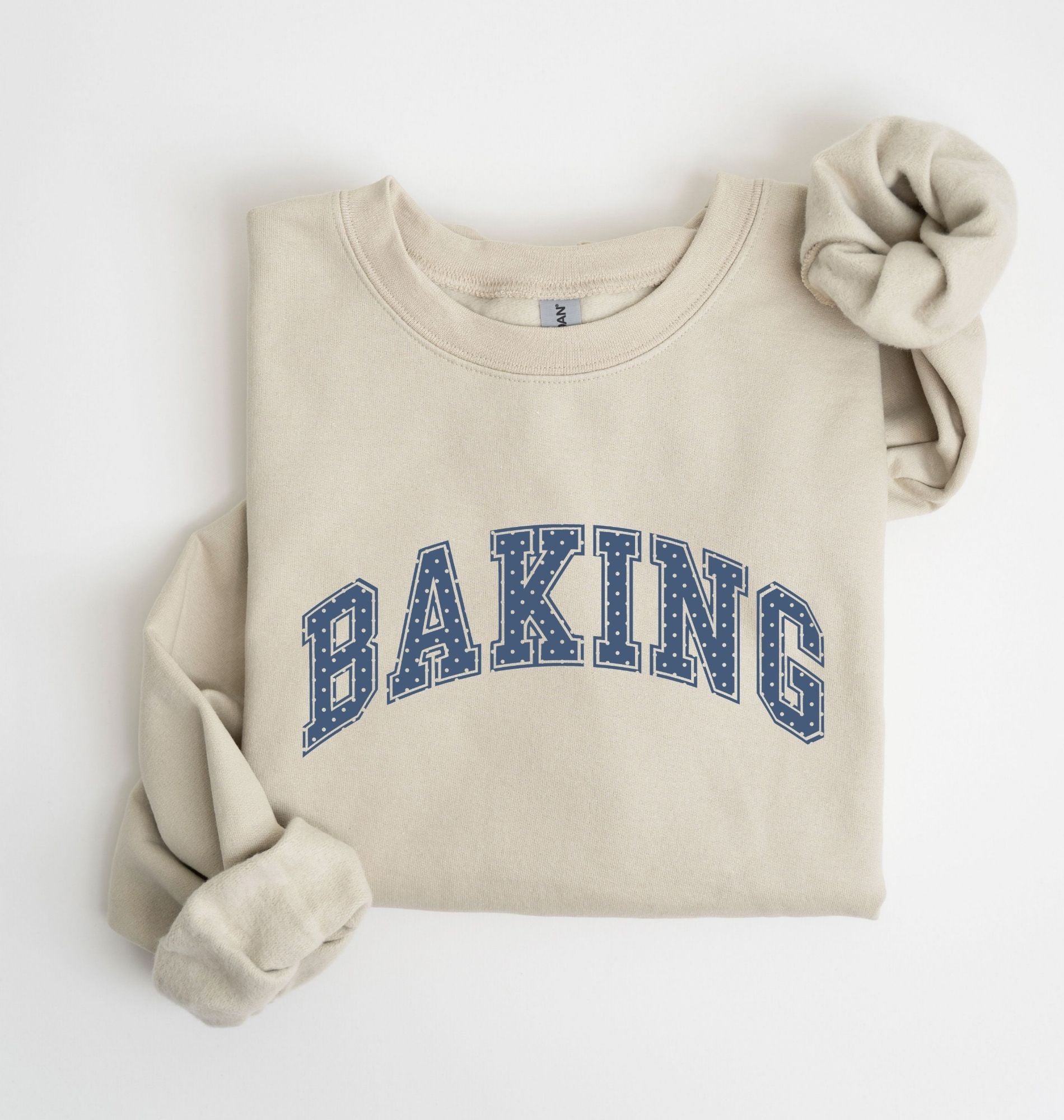 A folded cream-colored sweatshirt with 'BAKING' printed in blue varsity-style letters across the chest, styled with rolled sleeves on a white background.