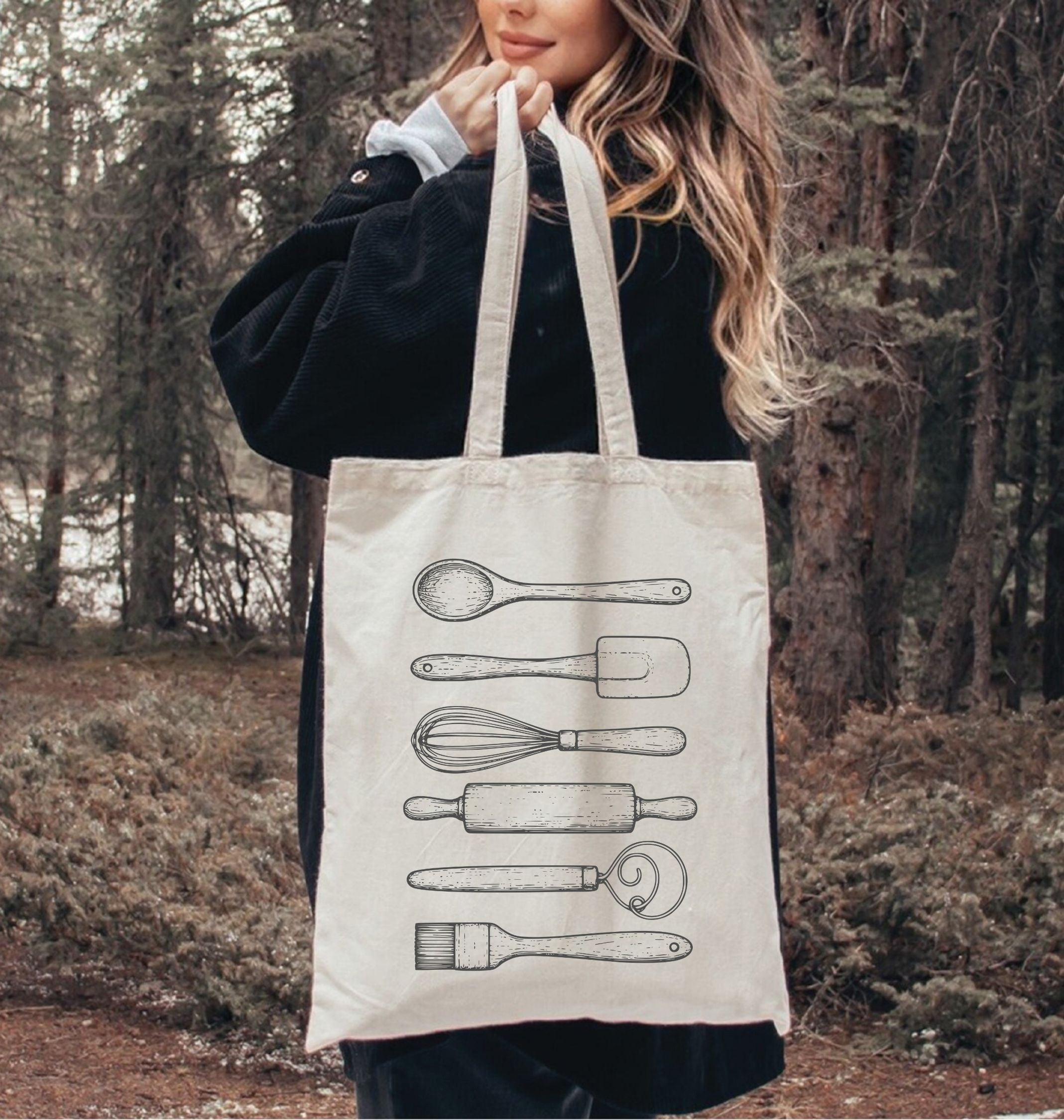 Person holding a tote bag with kitchen utensil illustrations in a forest setting