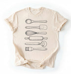 Flat ay of a Vintage baking tools shirt with whisk rolling pin and spatula illustrations on cream tee on a white background 