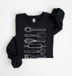 Flat lay of a black Vintage baking tools sweatshirt featuring whisk rolling pin spatula and kitchen utensil illustrations on a white background 