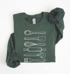 Flat lay of a forest green Vintage baking tools sweatshirt featuring whisk rolling pin spatula and kitchen utensil illustrations on a white background 
