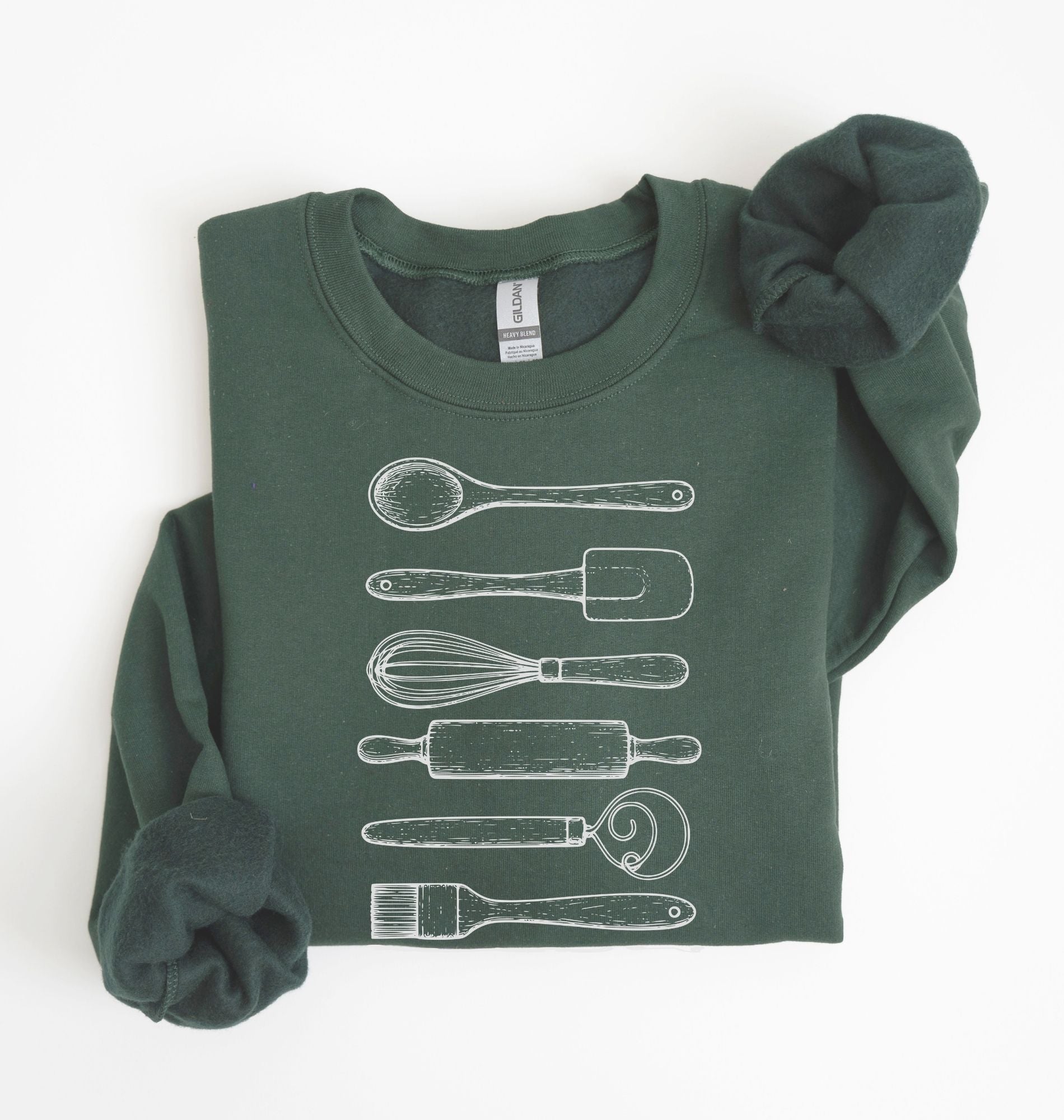 Flat lay of a forest green Vintage baking tools sweatshirt featuring whisk rolling pin spatula and kitchen utensil illustrations on a white background 