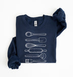 Flat lay of a navy blue Vintage baking tools sweatshirt featuring whisk rolling pin spatula and kitchen utensil illustrations on a white background 