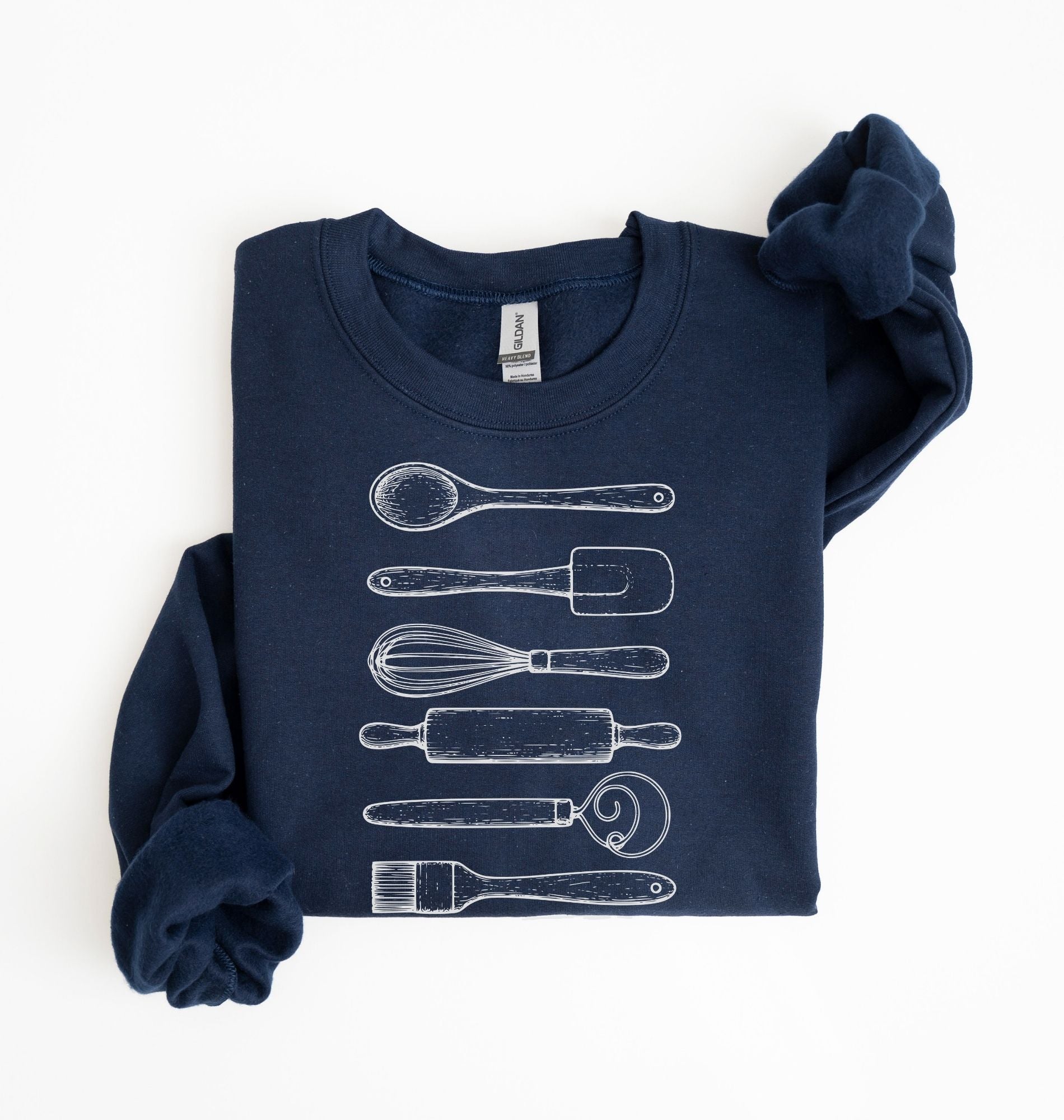 Flat lay of a navy blue Vintage baking tools sweatshirt featuring whisk rolling pin spatula and kitchen utensil illustrations on a white background 