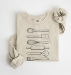 Flat lay of a sand Vintage baking tools sweatshirt featuring whisk rolling pin spatula and kitchen utensil illustrations on a white background 